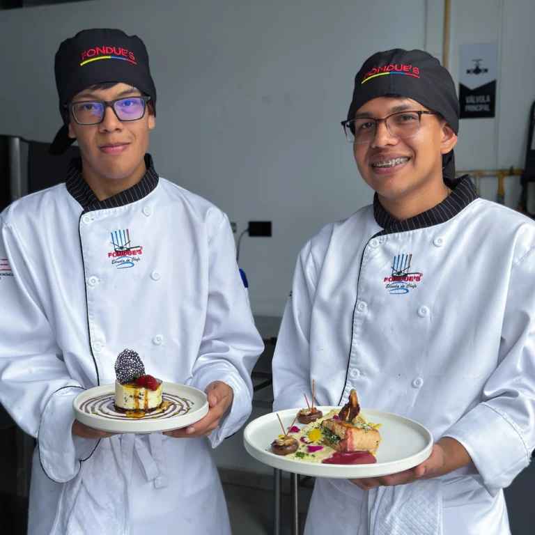 Dos estudiantes de la Escuela de Chefs Fondue's sonriendo en uniforme blanco, sosteniendo platos con postres de alta cocina presentados de forma artística.