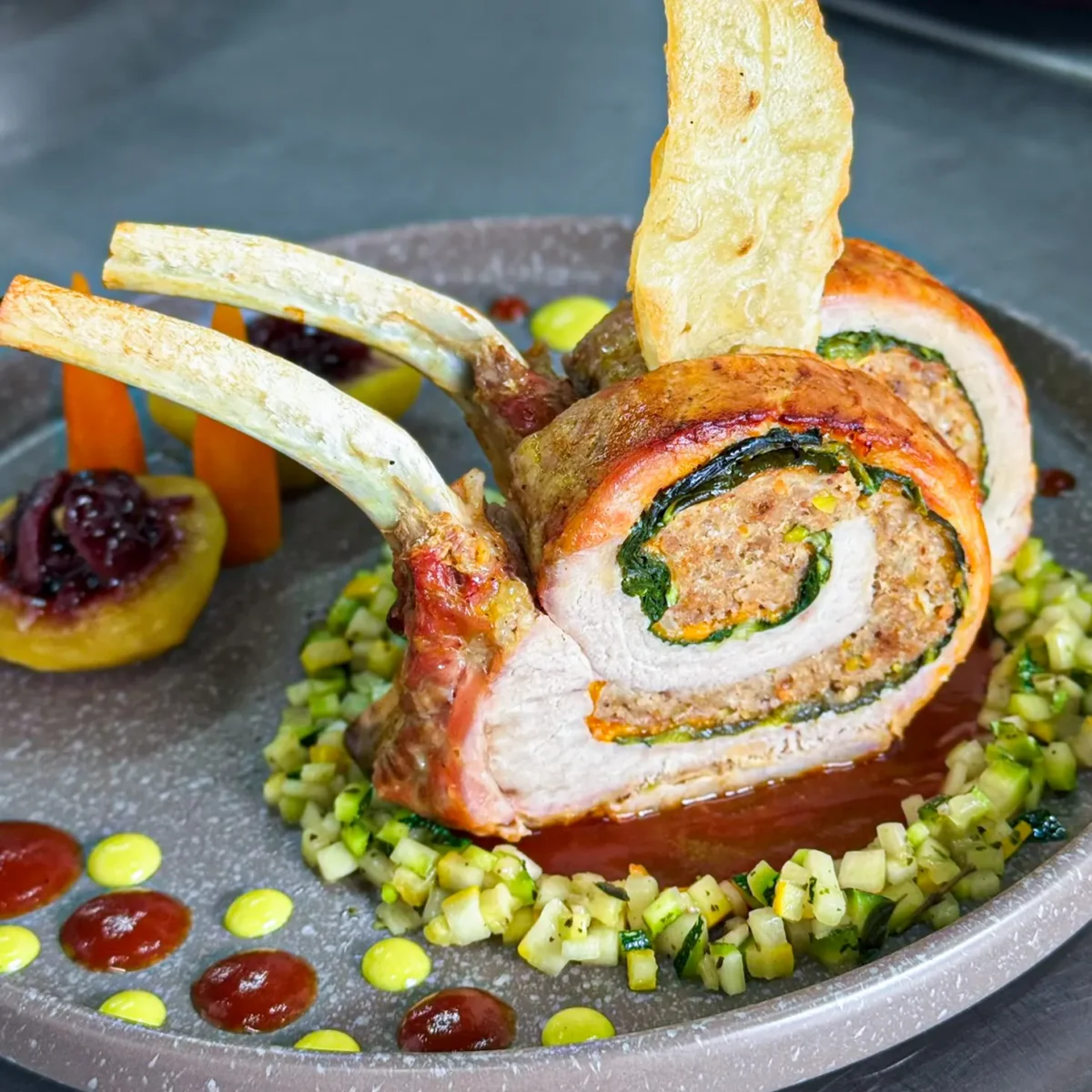 Plato gourmet de costillar de cordero relleno con espinacas, servido sobre una base de verduras picadas finamente y salsas decorativas.