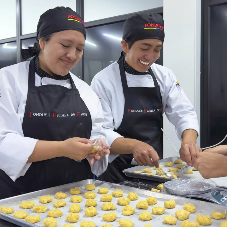 Dos estudiantes sonrientes de Fondue's Escuela de Chefs con delantales negros, preparando hileras de galletas sobre una bandeja para hornear.