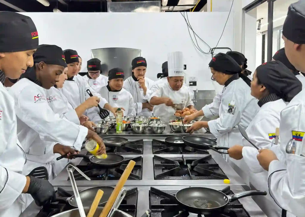 Grupo de estudiantes de gastronomía rodeando una estación de cocina industrial con sartenes y ollas mientras atienden la explicación de un instructor.