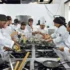 Grupo de estudiantes de gastronomía rodeando una estación de cocina industrial con sartenes y ollas mientras atienden la explicación de un instructor.